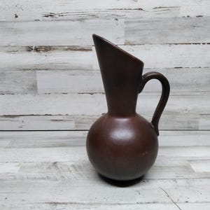 May include: A brown ceramic jug with a wide, flared spout and a handle. The jug has a rounded body and a smooth, matte finish.