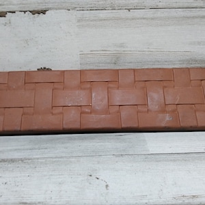 Puede incluir: Una caja rectangular de color marrón tejida con una tapa con bisagras. La caja está hecha de un material que se asemeja a la terracota.