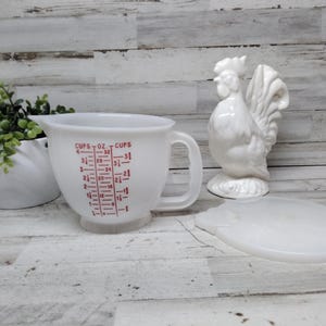 Könnte beinhalten: Weißer Messbecher mit roten Markierungen, einem Griff und einem Ausguss. Eine weiße Keramik-Hahn-Figur und ein weißer Deckel sind ebenfalls vorhanden. Ein kleiner weißer Pflanztopf mit Grünzeug befindet sich links.
