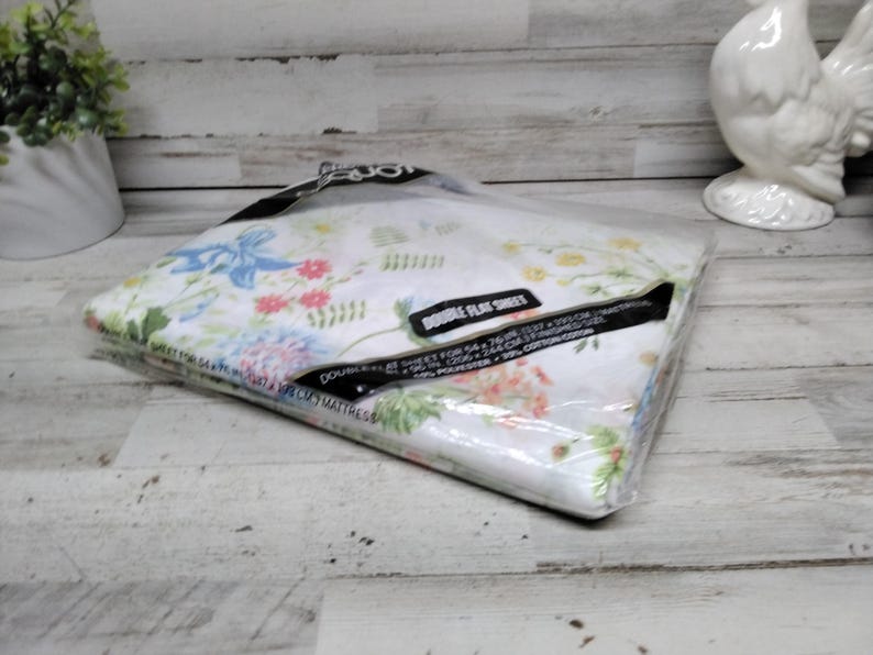 K&ouml;nnte beinhalten: Ein verpacktes Doppel-Flachbettlaken-Set mit floralem Muster in einer transparenten Plastikt&uuml;te. Das Bettw&auml;sche-Set hat einen wei&szlig;en Hintergrund mit bunten Blumen und blauen Schleifen. Die Verpackung ist mit "DOUBLE FLAT SHEET" beschriftet.