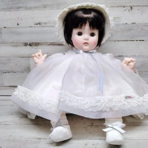 May include: A vintage porcelain doll with black hair, rosy cheeks, and a white bonnet. The doll wears a white dress with lace trim and a blue ribbon. White shoes and socks complete the outfit. The doll is a collectible item.
