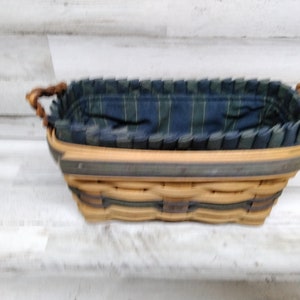 May include: Rectangular woven basket with a blue and green plaid fabric lining. The basket has a light brown exterior with a dark green trim and twisted handles. The interior fabric is pleated around the rim.