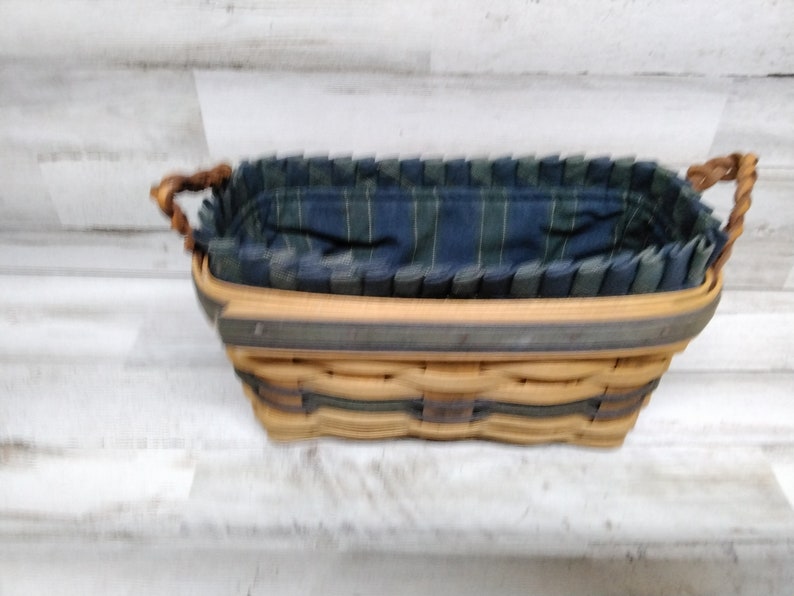 May include: Rectangular woven basket with a blue and green plaid fabric lining. The basket has a light brown exterior with a dark green trim and twisted handles. The interior fabric is pleated around the rim.