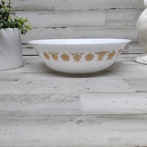 Puede incluir: Cuenco de cerámica blanco con un estampado floral dorado. El cuenco es redondo con un borde ligeramente ensanchado. El diseño floral se repite alrededor del exterior del cuenco. Un gallo decorativo de cerámica blanca y una maceta blanca están en el fondo.