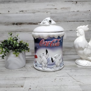 Op de afbeelding: Een witte keramische Coca-Cola koektrommel met een ijsberenontwerp, een witte keramische haanfiguur en een kleine witte keramische plantenbak met groen. De koektrommel heeft een deksel met een ijsbeer erop.