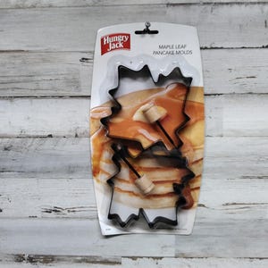 May include: Hungry Jack Maple Leaf Pancake Molds, a set of two black metal molds shaped like maple leaves, with wooden handles. The molds are shown on a white background with a stack of pancakes in the shape of maple leaves.