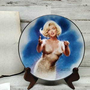 Puede incluir: Un plato de porcelana blanca con fondo azul que presenta un retrato de Marilyn Monroe con un vestido dorado brillante. El plato está sobre un soporte de madera y tiene la firma "Gene Autry" en la parte inferior.