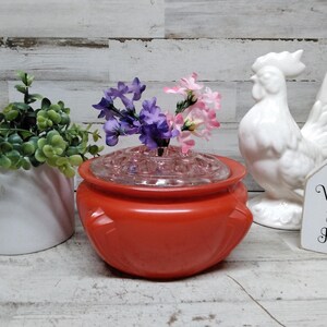 May include: A bright orange glass flower frog bowl with a clear glass lid featuring multiple holes for stems. The bowl contains small purple and pink artificial flowers. Beside it sits a white ceramic rooster figurine and a white planter.