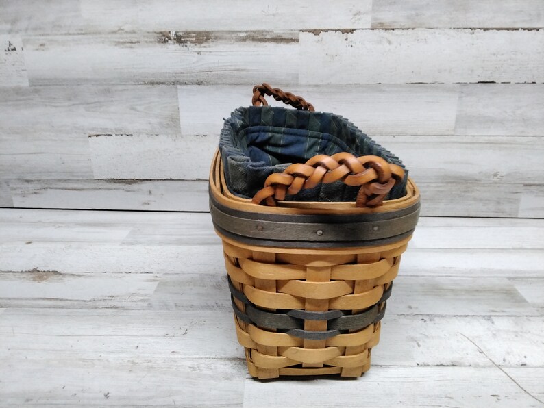 May include: A woven basket with a light brown body, dark grey bands and a braided leather handle. The interior has a blue and green plaid fabric lining. The basket has a square base and a rounded top, suitable for storage.