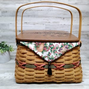 Può includere: Un cesto intrecciato beige con coperchio e manico in legno. Il cesto ha dettagli rossi e verdi. Un tessuto fantasia con un disegno di frutta e fiori copre la parte superiore. Una nappa nera pende dalla parte anteriore.