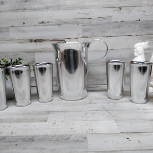 May include: A set of polished silver drinkware, including a pitcher with a handle and six matching tumblers. The reflective surfaces create a bright, clean aesthetic. The pitcher is approximately 9 inches tall.