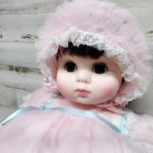 Puede incluir: Una muñeca vintage con un vestido rosa y un gorro a juego. La muñeca tiene cabello oscuro, ojos grandes y mejillas rosadas. El gorro está adornado con encaje y tul, y el vestido tiene una cinta de satén.