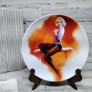 Puede incluir: Un plato de cerámica blanco con una imagen colorida de una mujer con un suéter morado y medias negras sentada en un taburete. El fondo es un degradado de naranja y amarillo.