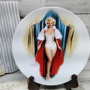 Puede incluir: Un plato de cerámica blanco con una imagen colorida de una mujer con un vestido blanco y una capa roja. La mujer está de pie frente a una cortina azul y dorada. El plato está sobre un soporte de madera.