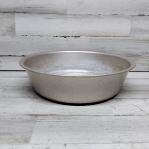 Op de afbeelding: Een ronde, zilverkleurige metalen kom met een breed, ondiep ontwerp. De kom heeft een licht versleten uiterlijk, wat suggereert dat hij vintage of veel gebruikt kan zijn. De rand is gebogen en de basis is plat.