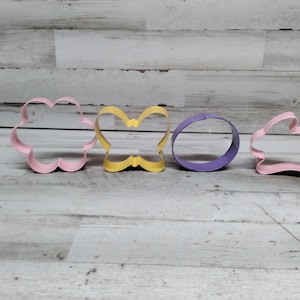 Puede incluir: Cuatro cortadores de galletas de metal en diferentes formas: una flor, una mariposa, un círculo y un conejito. Los cortadores de flores y conejitos son rosas, el cortador de mariposas es amarillo y el cortador de círculos es morado.