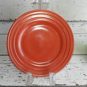 May include: A round, orange-red ceramic plate with three concentric rings near the rim. The plate is displayed on a clear, acrylic stand against a white, distressed wood background. A green glass bottle is partially visible.