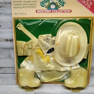 May include: A Cabbage Patch Kids Western Collection costume set for children aged 3 and over. The set includes a cream-coloured cowboy hat, shirt, belt, and boots. The packaging features text that reads "turn into cowgirls and cowboys when they're wearing WESTERNS."