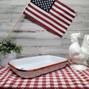 Könnte beinhalten: Eine rechteckige Backform aus weißem Emaille mit rotem Rand liegt auf einem rot-weiß karierten Tuch. Eine weiße Keramik-Hahnfigur und eine kleine amerikanische Flagge sind ebenfalls vorhanden. Eine kleine Topfpflanze ist ebenfalls im Bild.