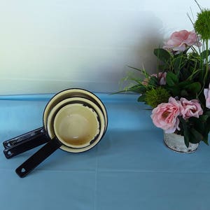 Puede incluir: Un juego de tres tazas medidoras apilables con mangos negros e interior color crema. Las tazas están dispuestas sobre una superficie azul claro, con un arreglo floral de rosas rosas y vegetación en el fondo.