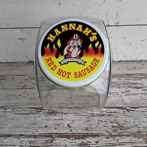 以下が含まれることがあります： 「HANNAH'S RED HOT SAUSAGE」の文字が入った白い蓋が付いた、透明な長方形のガラス瓶。 蓋には、消防士の衣装を着た漫画の豚が炎に囲まれて描かれています。 瓶は、使い古された白い木製の表面に置かれています。
