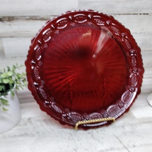 May include: A ruby red glass plate with a decorative scalloped edge. The plate is round and has a textured, radial pattern in the center. It is displayed on a gold-colored metal stand. A small potted plant and a white ceramic chicken are in the background.