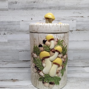 Puede incluir: Un tarro de cerámica blanco con una tapa de hongo. El tarro está decorado con un patrón de hongos y hojas en tonos de verde, marrón y amarillo.