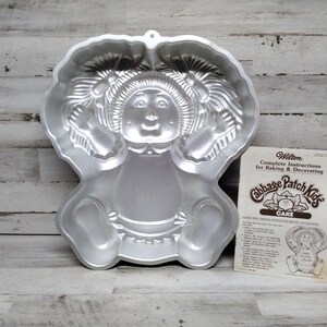 Puede incluir: Molde para pastel de metal plateado con forma de muñeca Cabbage Patch Kid. El molde presenta un diseño detallado de una muñeca con coletas, un vestido y zapatos. El molde viene con una hoja de instrucciones impresa para hornear y decorar un pastel Cabbage Patch Kid.