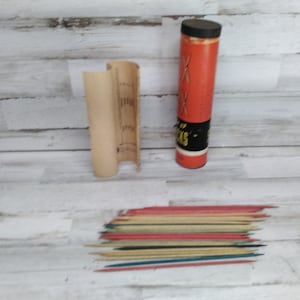 Peut inclure: Un jeu vintage avec un tube rouge et noir, le mot "STICKS" et un couvercle noir. Une pochette en papier beige et un tas de bâtonnets colorés sont également présents. Les bâtonnets sont rouges, bleus, jaunes et noirs.