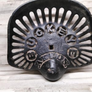 Könnte beinhalten: Schwarzer gusseiserner Traktorsitz mit geschlitzter Rückenlehne und dem Wort "AKRON" auf der Vorderseite. Der Sitz hat einen abgerundeten Boden und die Buchstaben "G O K E Y E" in der Mitte.