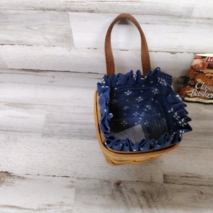 May include: A small, blue and white patterned basket with a brown handle. The basket is made of woven wicker and has a fabric lining. The basket is sitting on a white wooden surface.