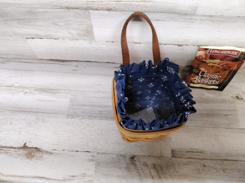 May include: A small, blue and white patterned basket with a brown handle. The basket is made of woven wicker and has a fabric lining. The basket is sitting on a white wooden surface.