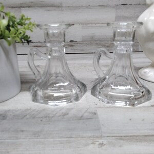 May include: Two clear glass candle holders with handles. The candle holders have a hexagonal base and a flared body. The handles are curved and attached to the side of the candle holder. The candle holders are on a white wooden surface.