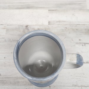 May include: A white ceramic mug with a blue stripe around the rim and handle. The mug is empty and sitting on a white wooden surface.
