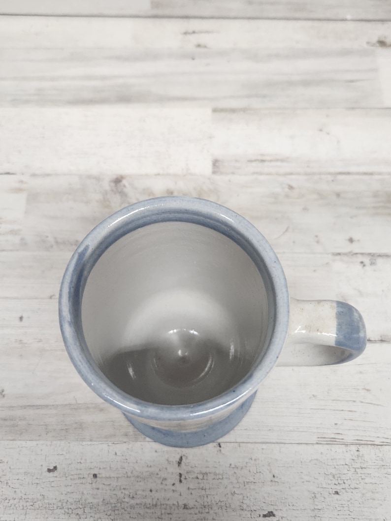 May include: A white ceramic mug with a blue stripe around the rim and handle. The mug is empty and sitting on a white wooden surface.