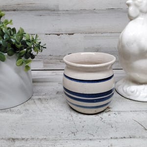 May include: A small, off-white ceramic pot with horizontal blue stripes. The pot is next to a white ceramic rooster figurine and a white planter with green foliage. The objects are on a distressed, white wooden surface.