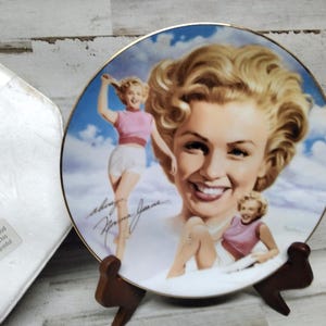 Puede incluir: Un plato de cerámica blanco con borde dorado presenta una imagen colorida de Marilyn Monroe. El plato está sobre un soporte de madera. El texto "Marilyn Monroe" está escrito con tinta negra en el plato.