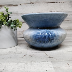 May include: A light blue ceramic planter with a flared rim and a textured basket-weave pattern. The exterior features a raised, embossed floral design in a darker blue hue. The bowl-shaped vessel has a wide opening and a rounded base.