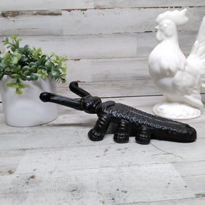 May include: A black cast iron bottle opener in the shape of an alligator. A white ceramic rooster and a white ceramic pot with green leafy plants are also in the image.