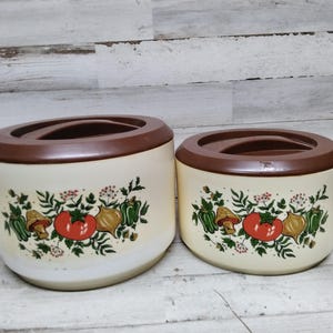 May include: Two vintage kitchen canisters with brown lids and white bodies. The canisters have a floral design with red tomatoes, green peppers, and brown mushrooms.