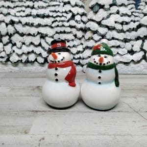 May include: Two ceramic snowman figurines. One snowman wears a black top hat and red scarf, the other wears a green hat and scarf. Both have orange carrot noses, black button eyes, and a black button mouth. The snowmen are on a white and grey wooden surface.