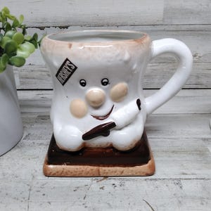 Może przedstawiać: Kapryśny ceramiczny kubek w kształcie s'more, z twarzą pianki marshmallow i logo Hershey's. Kubek ma brązową podstawę i biały uchwyt, idealny do gorących napojów.