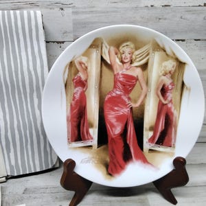 Puede incluir: Un plato de cerámica blanco con una imagen roja y blanca de una mujer con un vestido rojo. El plato está sobre un soporte de madera. La imagen es de una mujer con un vestido rojo de pie frente a un espejo. La mujer se está mirando en el espejo. El plato es un artículo de colección.