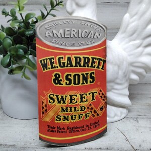 Puede incluir: Una lata de rapé Sweet Mild Snuff de W.E. Garrett & Sons, de estilo vintage. La lata es roja con texto dorado y negro, incluyendo "AMERICAN SINCE 1782". La parte inferior de la lata dice "Trade Mark Registered in United States Patent Office, Oct. 25. 1870".