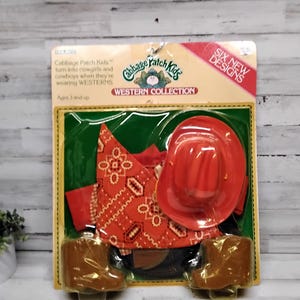 May include: Cabbage Patch Kids Western Collection playset. The set includes a red cowboy hat, a red bandana with a white pattern, a red shirt, and brown boots. The packaging is yellow and green.
