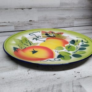 Peut inclure: Une assiette en &eacute;mail vintage avec un bord vert et un centre blanc. L'assiette pr&eacute;sente un motif peint &agrave; la main de fruits, y compris des pommes et des raisins, dans des tons de jaune, orange, rouge et vert.
