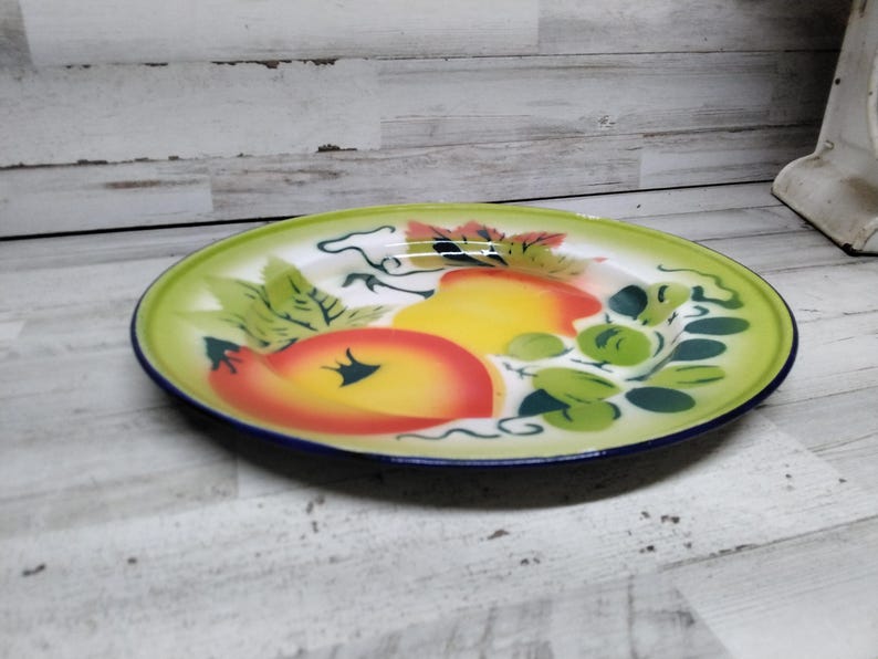 Peut inclure: Une assiette en &eacute;mail vintage avec un bord vert et un centre blanc. L'assiette pr&eacute;sente un motif peint &agrave; la main de fruits, y compris des pommes et des raisins, dans des tons de jaune, orange, rouge et vert.