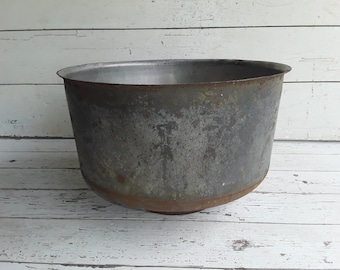 May include: A large, weathered metal bowl with a wide, open top and a rounded base. The bowl has a dark, mottled gray finish with areas of rust, suggesting age and use. The bowl is likely used for cooking or storage.