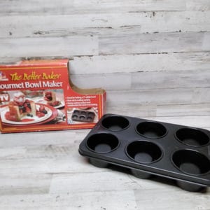 May include: A red and white box with the text "The Better Baker Gourmet Bowl Maker" and a picture of a dessert. A black metal baking pan with six round cavities.