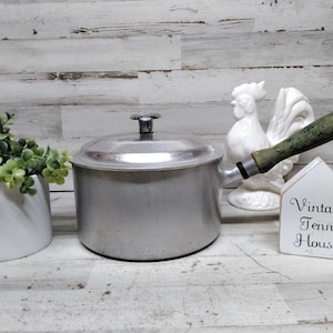 May include: A silver aluminum saucepan with a lid and a long, weathered green wooden handle. Next to the pan is a white ceramic rooster figurine and a small white house-shaped sign with the black text Vintage Tenn House printed on the front.